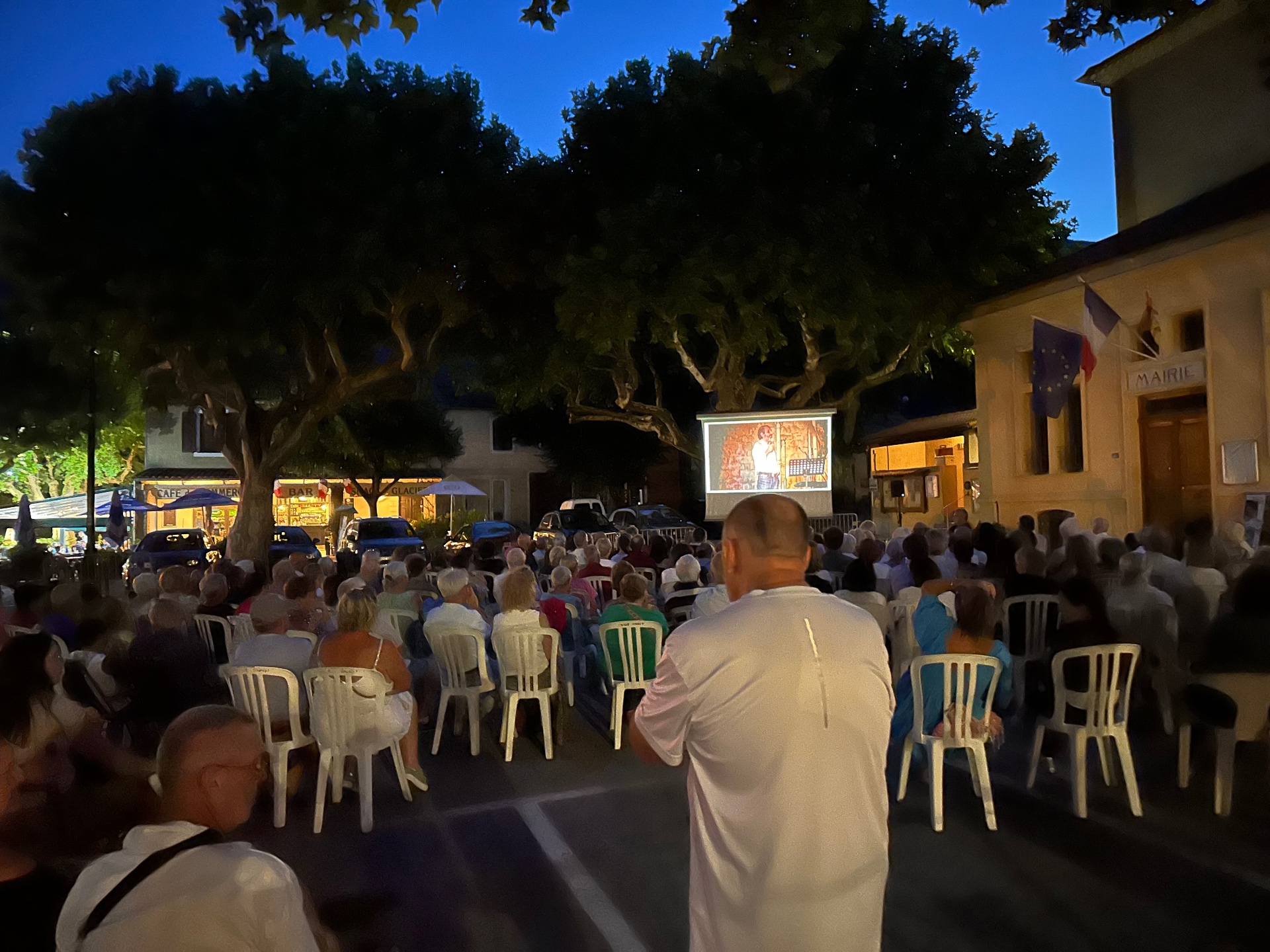 Projection en plein air à la tombée de la nuit place de la Mairie à Annot, l'écran et le public sous les platanes.